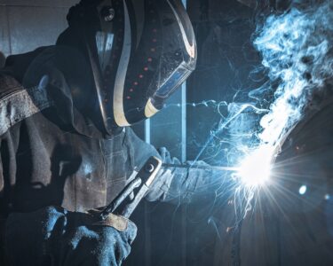 A man wearing a helmet and a fire-resistant bodysuit uses a welding torch on a piece of metal, sending out sparks and smoke.