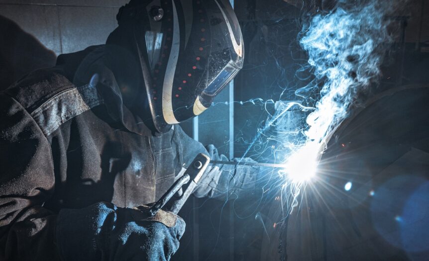 A man wearing a helmet and a fire-resistant bodysuit uses a welding torch on a piece of metal, sending out sparks and smoke.