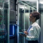 A technician wearing a white blouse and holding a tablet looks at a row of lit up server racks behind glass doors.