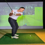 A young man is golfing indoors using a golf simulator game. He's wearing golfing pants, shirts, and shoes.