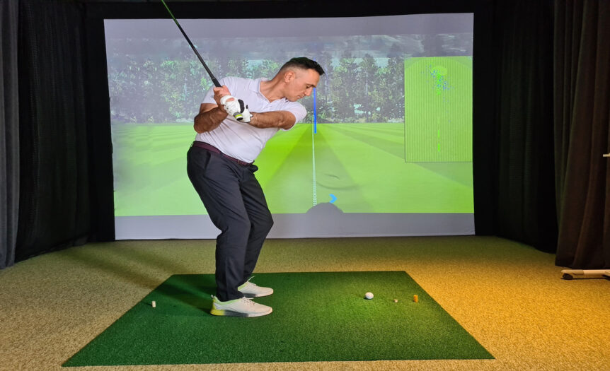 A young man is golfing indoors using a golf simulator game. He's wearing golfing pants, shirts, and shoes.