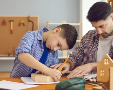 A father and son work together on a birdhouse. The child draws on one side of the house with a pencil.