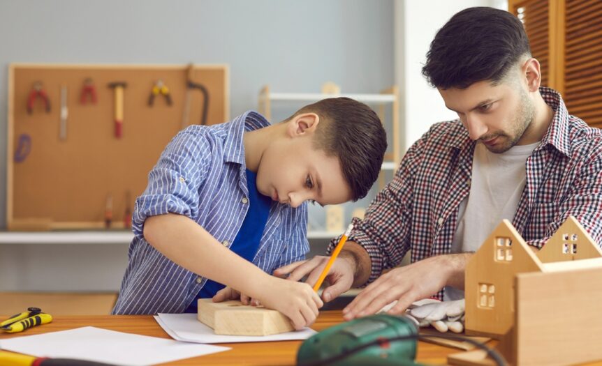 A father and son work together on a birdhouse. The child draws on one side of the house with a pencil.