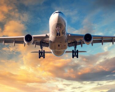 A large, white airplane is flying against a blue and yellow cloud-filled sky. It has its landing gear out.
