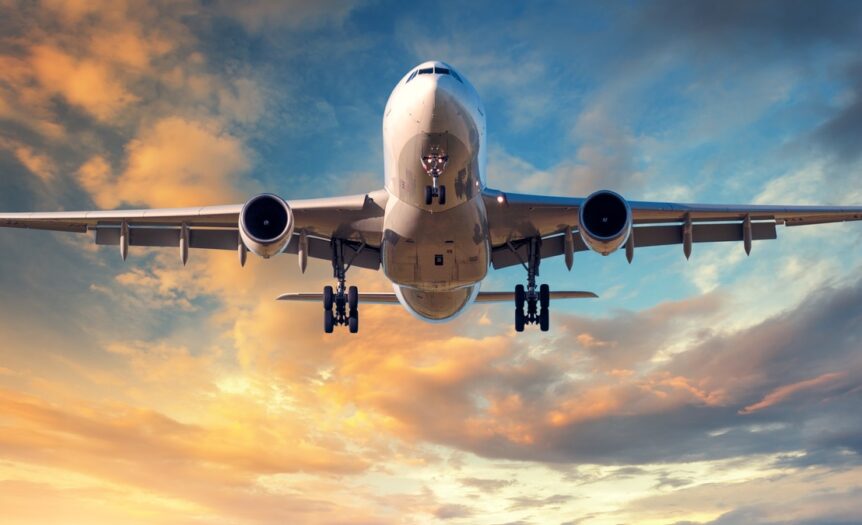 A large, white airplane is flying against a blue and yellow cloud-filled sky. It has its landing gear out.