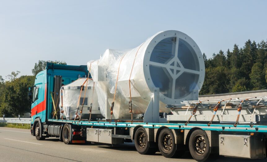 A blue semi-truck hauling a white, disassembled wind turbine on a highway, secured with orange load binders.