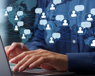 A man in a blue shirt types on a laptop at a desk, with a world map and icons showing global communication overlaid.