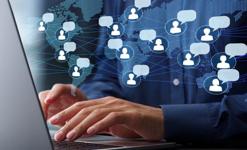 A man in a blue shirt types on a laptop at a desk, with a world map and icons showing global communication overlaid.