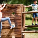A young girl smiles while climbing on a wooden vertical climber. A young boy and girl walk across wooden beams in the background.