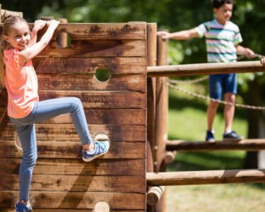 A young girl smiles while climbing on a wooden vertical climber. A young boy and girl walk across wooden beams in the background.