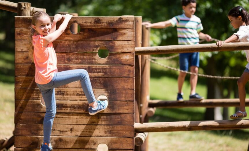 A young girl smiles while climbing on a wooden vertical climber. A young boy and girl walk across wooden beams in the background.