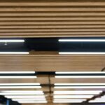 Overhead lighting strips in between wood spacers in an office space, with a screen on the wall in the background.