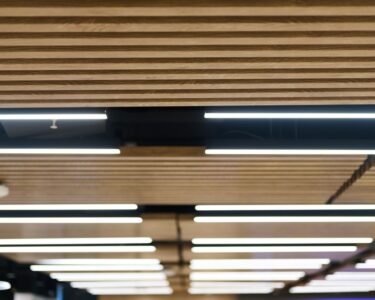 Overhead lighting strips in between wood spacers in an office space, with a screen on the wall in the background.
