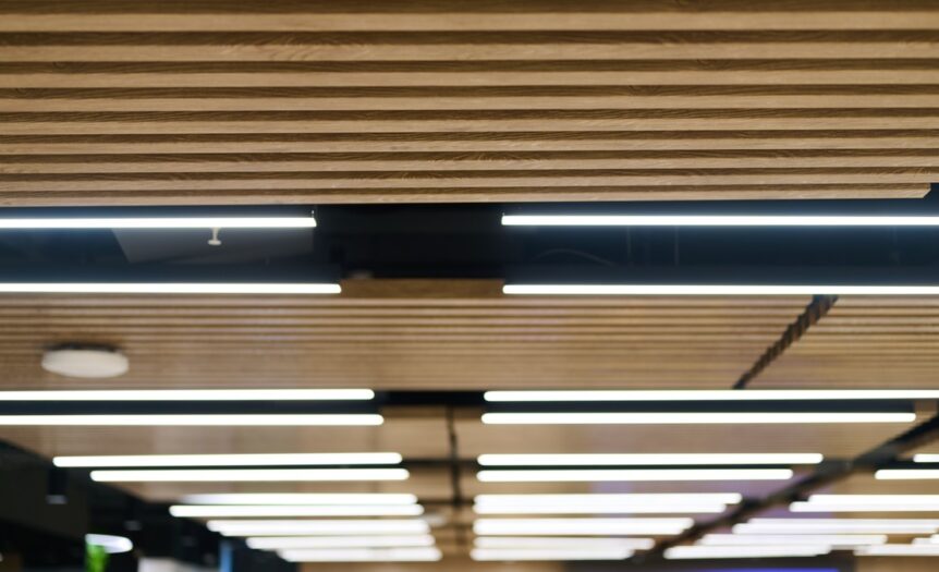 Overhead lighting strips in between wood spacers in an office space, with a screen on the wall in the background.