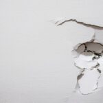 A white-painted drywall with a hole on the right side, with cracks spreading out from the top and bottom of the damaged area.