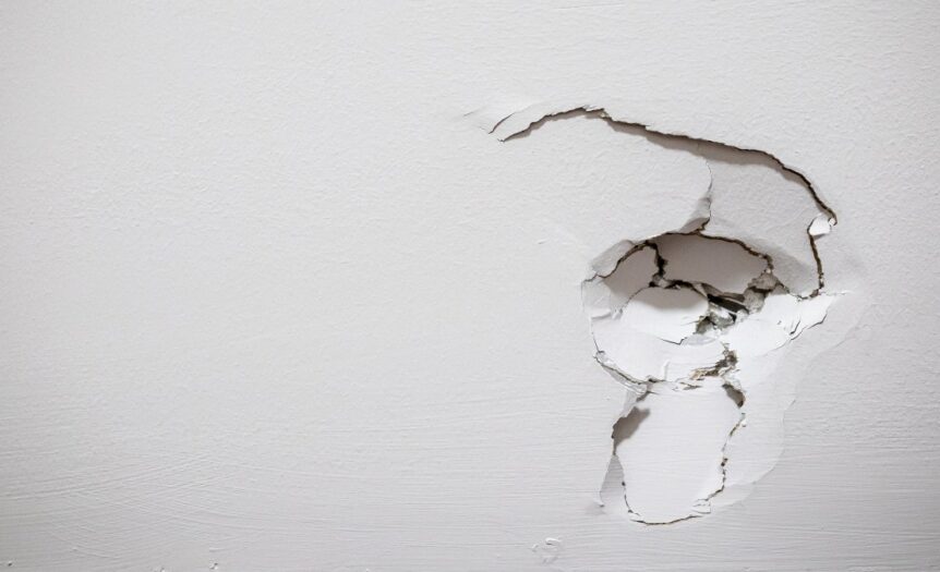 A white-painted drywall with a hole on the right side, with cracks spreading out from the top and bottom of the damaged area.