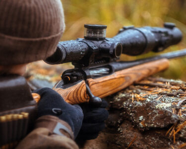 A man lies on his stomach in a beanie and jacket, steadying a wet rifle on timber while looking through the scope.