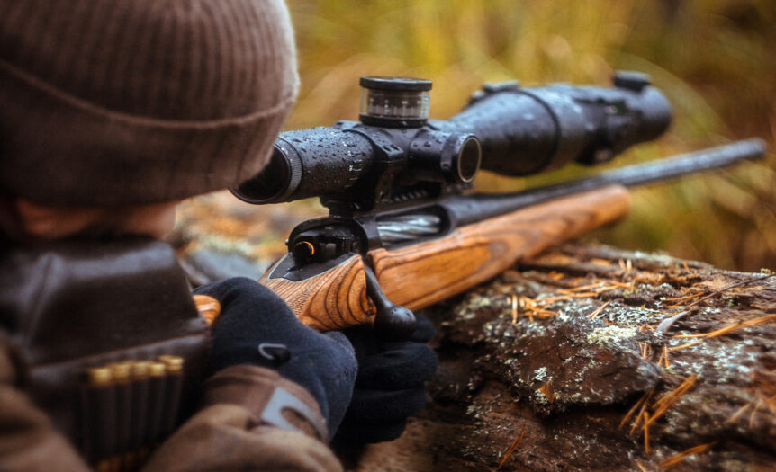 A man lies on his stomach in a beanie and jacket, steadying a wet rifle on timber while looking through the scope.