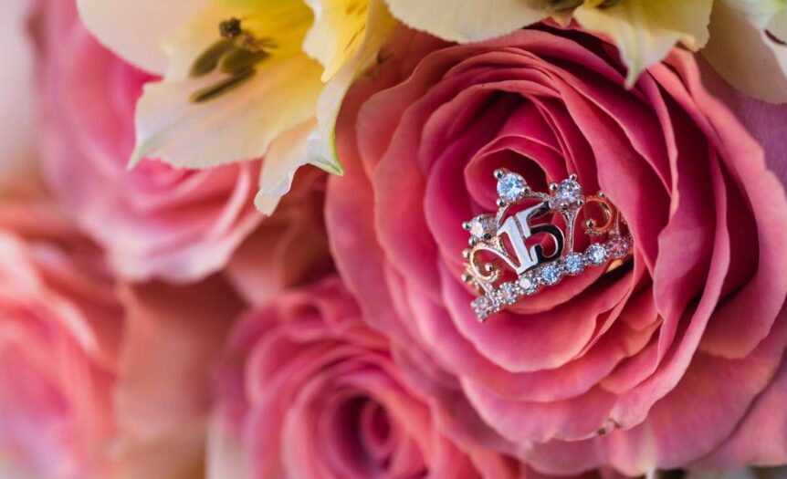 A pink rose bouquet with a silver "15" ring adorned with gemstones in the center; other roses and lilies surround it.