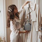 A woman in a white dress shirt and cream, high-waisted pants holds up a trench coat and a small, round purse.