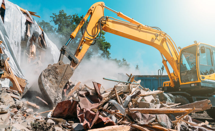 An excavator scoops up scraps of metal, dirt, and other building materials. Dust and debris fill the air.