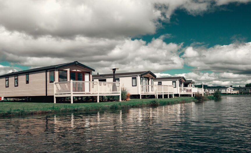 A row of white and tan mobile homes sits along the edge of a body of water. Each home has a porch.