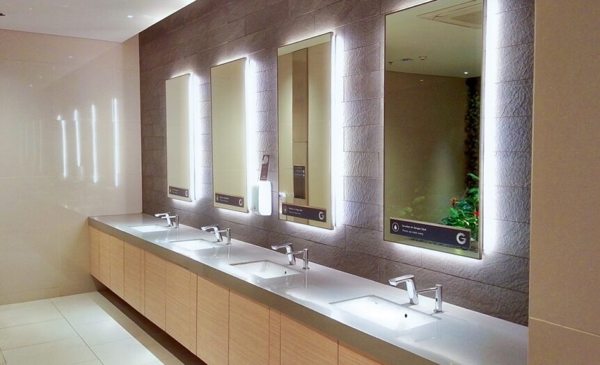 A commercial restroom has four sinks with gray countertops. Four mirrors have lighting behind them above the sinks.