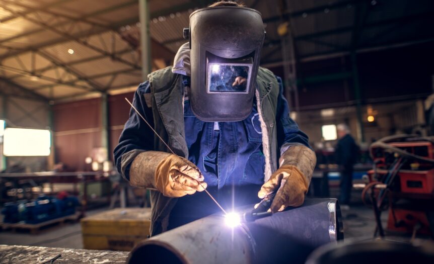 A person wearing a protective face mask and apron is using a welder against some metal. They are also wearing gloves.