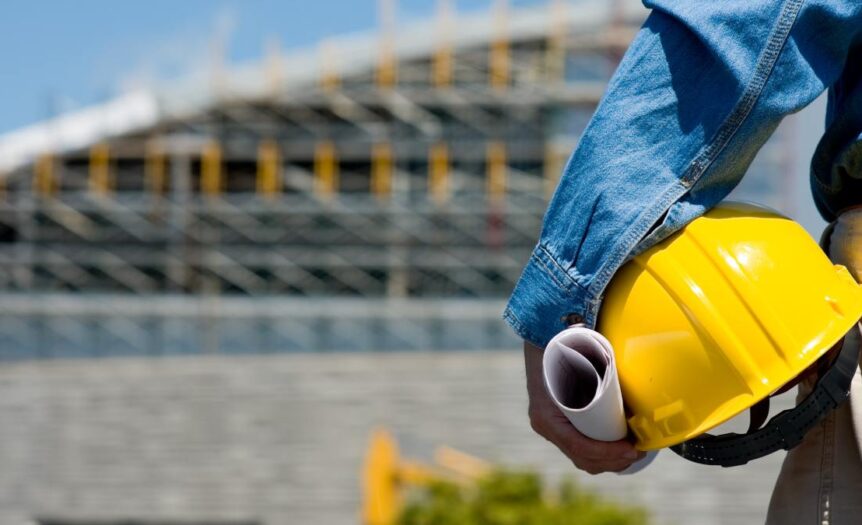 A person wearing a jean jacket holds a hard hat and paper on one hand. Behind is a building under construction.