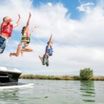 Three kids are having fun jumping off a speedboat in multi-colored floaties and life jackets as their mom watches.