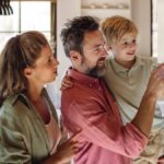 A woman and a man carrying a young boy looking at a large thermostat on the wall inside a dining room.