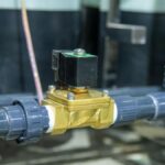 A brass solenoid valve with a black top sits between two gray, plastic pipes. A gray plastic pipe is behind that one.