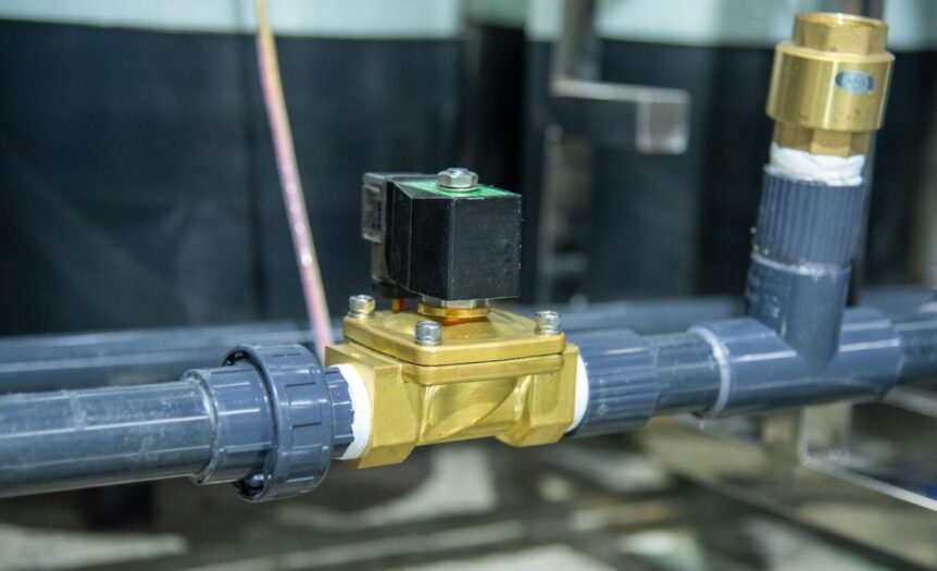 A brass solenoid valve with a black top sits between two gray, plastic pipes. A gray plastic pipe is behind that one.