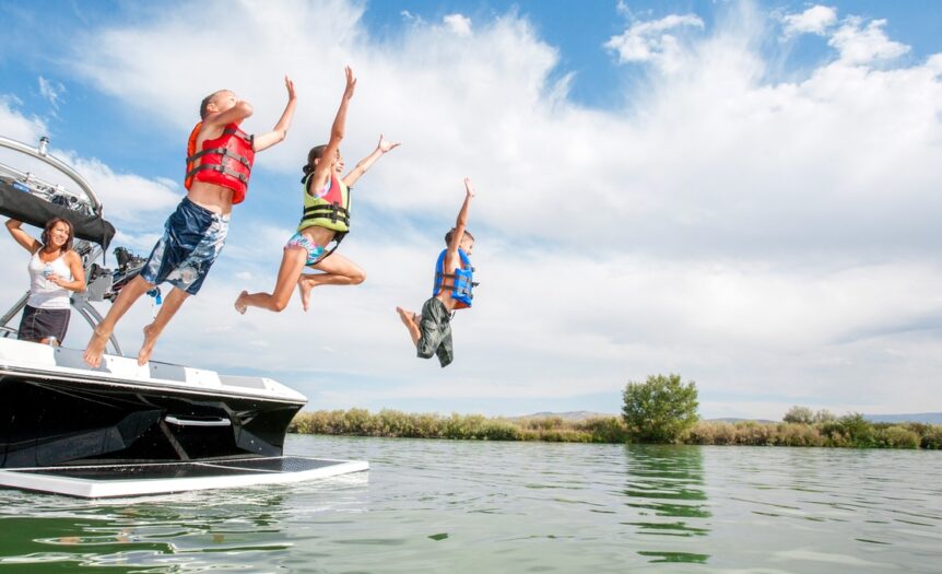 Kids in life jackets jump off the back of a boat into a lake with their mom standing in the boat watching them.