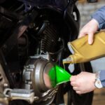 A person in a blue coat uses a green funnel to pour oil from a gold container into a motorcycle engine.