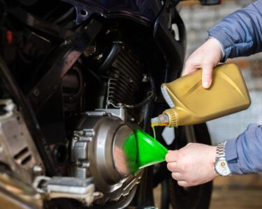 A person in a blue coat uses a green funnel to pour oil from a gold container into a motorcycle engine.