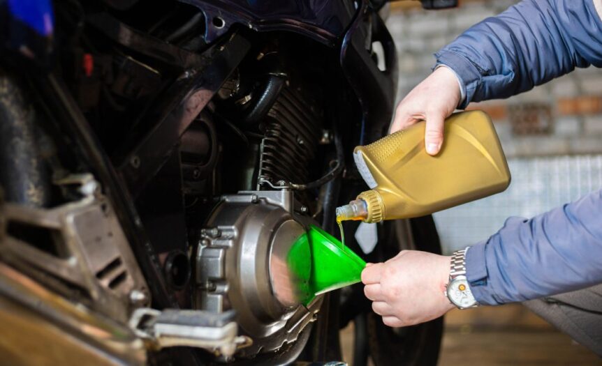 A person in a blue coat uses a green funnel to pour oil from a gold container into a motorcycle engine.
