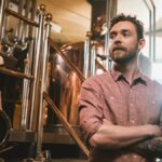A young man with tattoos stands with his arms crossed, with brass distillery equipment used for brewing behind him.