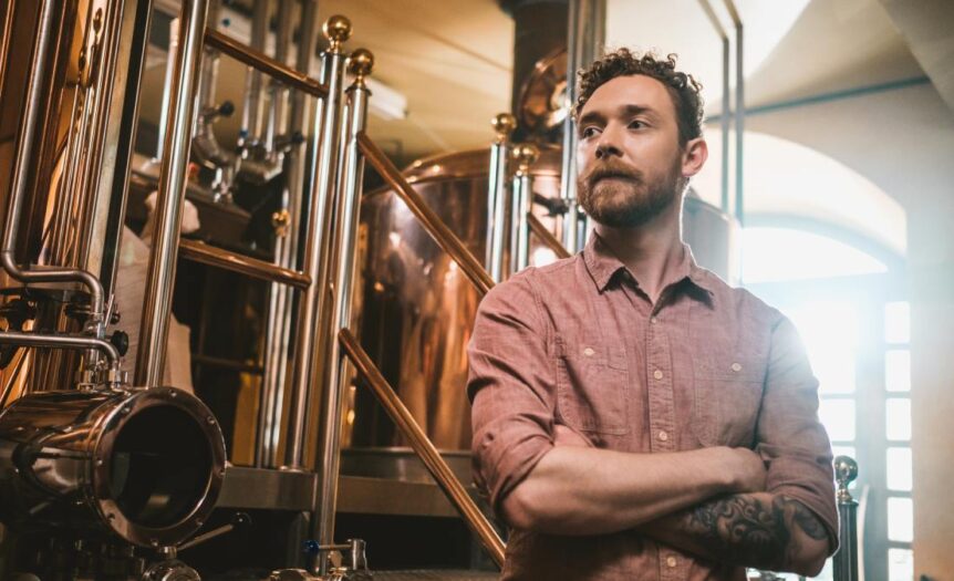 A young man with tattoos stands with his arms crossed, with brass distillery equipment used for brewing behind him.