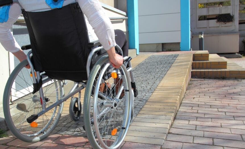 A person in a wheelchair is at the foot of a ramp. The ramp leads up onto a porch area. There are steps next to the ramp.