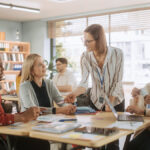A group of five elementary school teachers sitting and standing around a table during a faculty meeting.