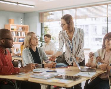 A group of five elementary school teachers sitting and standing around a table during a faculty meeting.