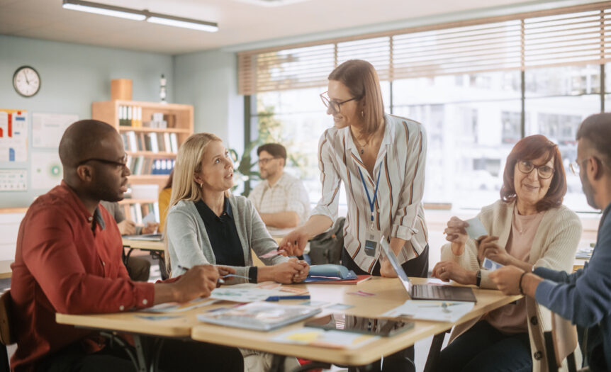 A group of five elementary school teachers sitting and standing around a table during a faculty meeting.