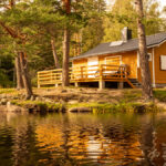 A small cabin in the woods sits on the edge of a lake. It has tan walls and a wooden deck in the back.