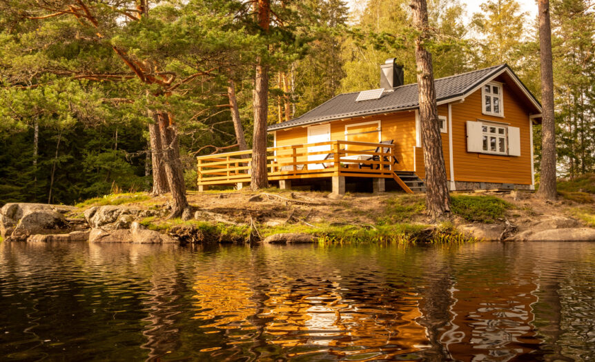 A small cabin in the woods sits on the edge of a lake. It has tan walls and a wooden deck in the back.