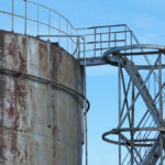 A side staircase leading up to an old, metal water container tank with rust marks against a blue sky.