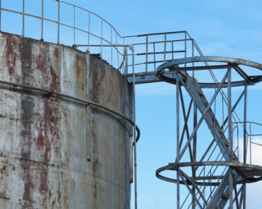 A side staircase leading up to an old, metal water container tank with rust marks against a blue sky.