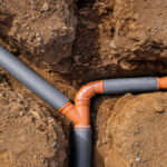 A collection of unfinished gray pipes with orange connectors arranged in narrow trenches in light brown soil.