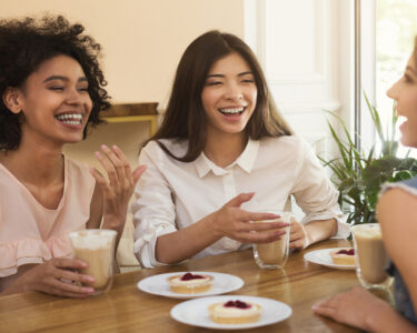Three women sitting together at a table by a window, enjoying coffees and tarts while chatting happily.