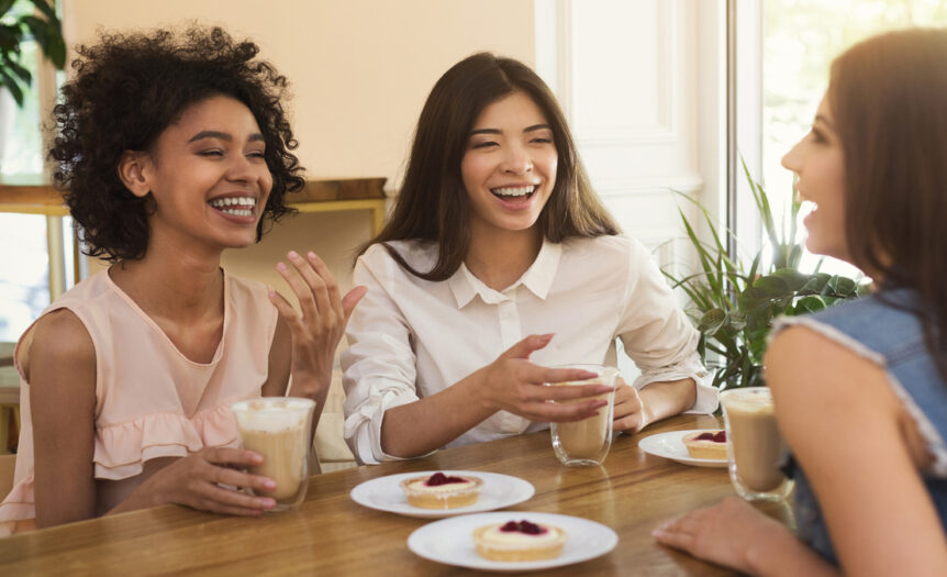 Three women sitting together at a table by a window, enjoying coffees and tarts while chatting happily.